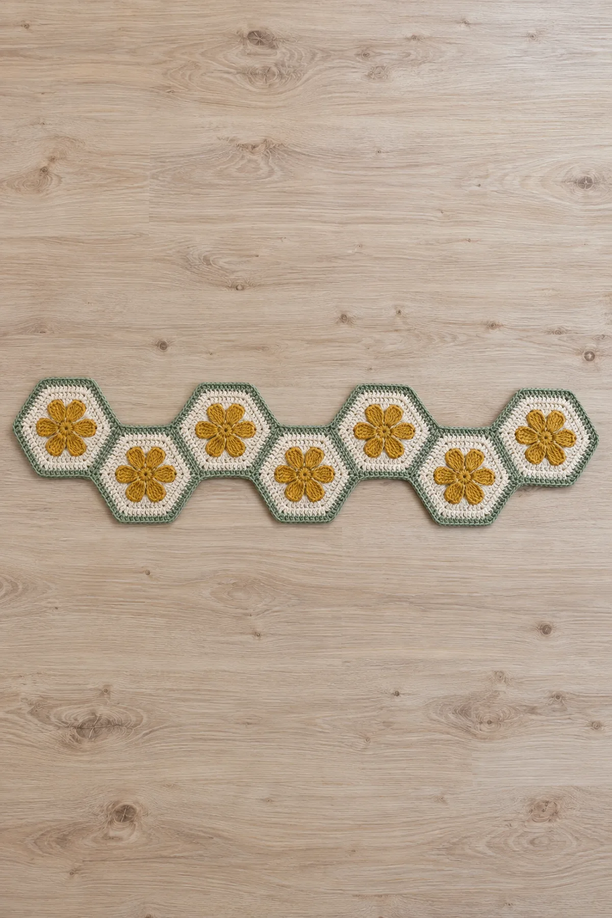 Original narrow crochet table runner of EXACTLY SEVEN cream hexagons each containing one flat mustard SIX-petal daisy, sage flat seams and tiny continuous sage sc border. Seven motifs joined in ONE gently staggered zigzag chain from left to right, not a double row or cluster. All hexagons flat-top oriented, alternating slightly down and up. Approx22inches long5inches wide. Entire runner visible on pale wood, no extra flowers, no props on runner