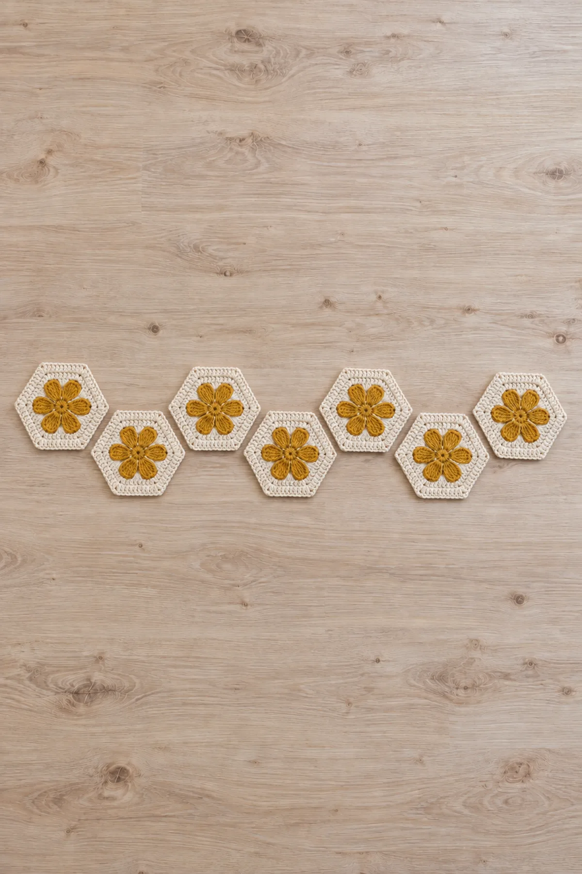 EXACTLY seven separate complete cream crochet hexagons with flat mustard six-petal daisies, arranged as one staggered zigzag chain on pale table BEFORE sage joining, visible gaps between all seven, no joined border