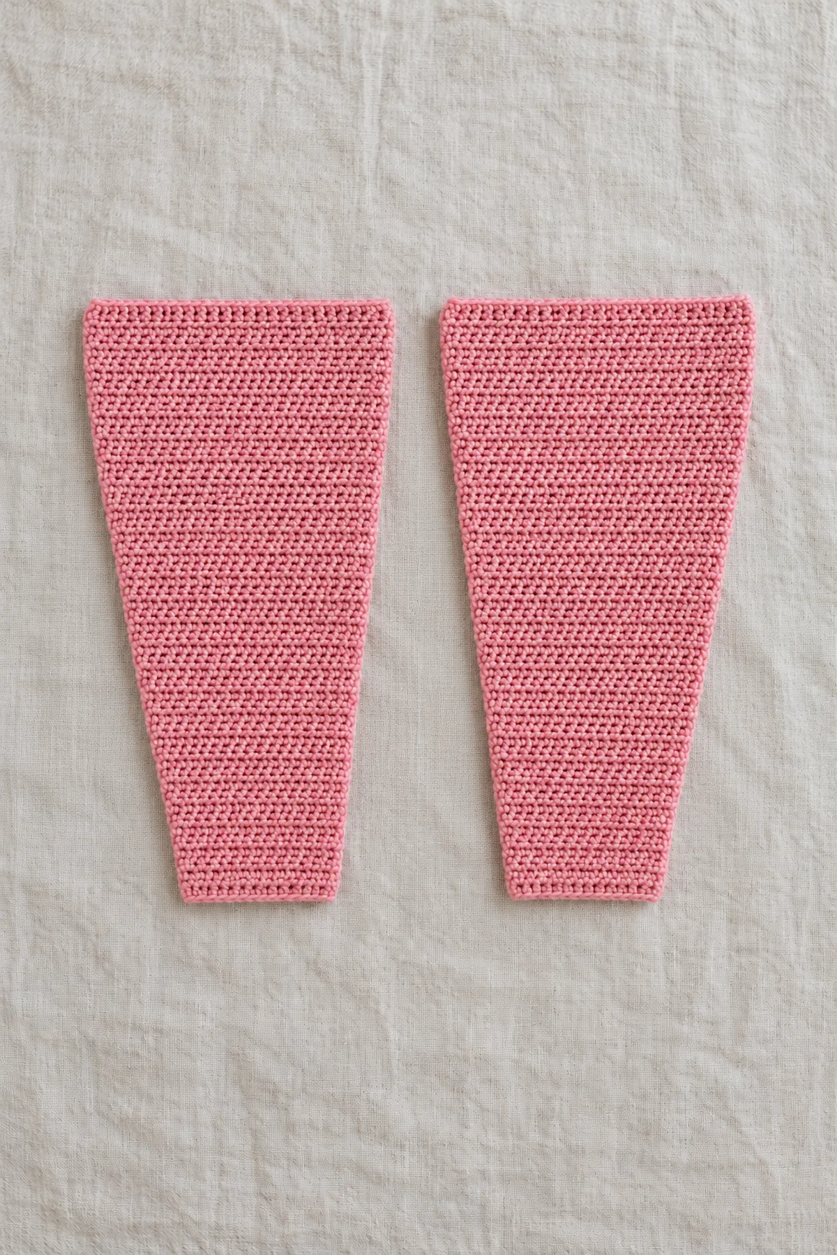 Two separate gently tapered rose-pink crochet sleeve panels, wide straight upper edges and narrower cuffs, plus the three straight body panels nearby, all unsewn; no finished garment.