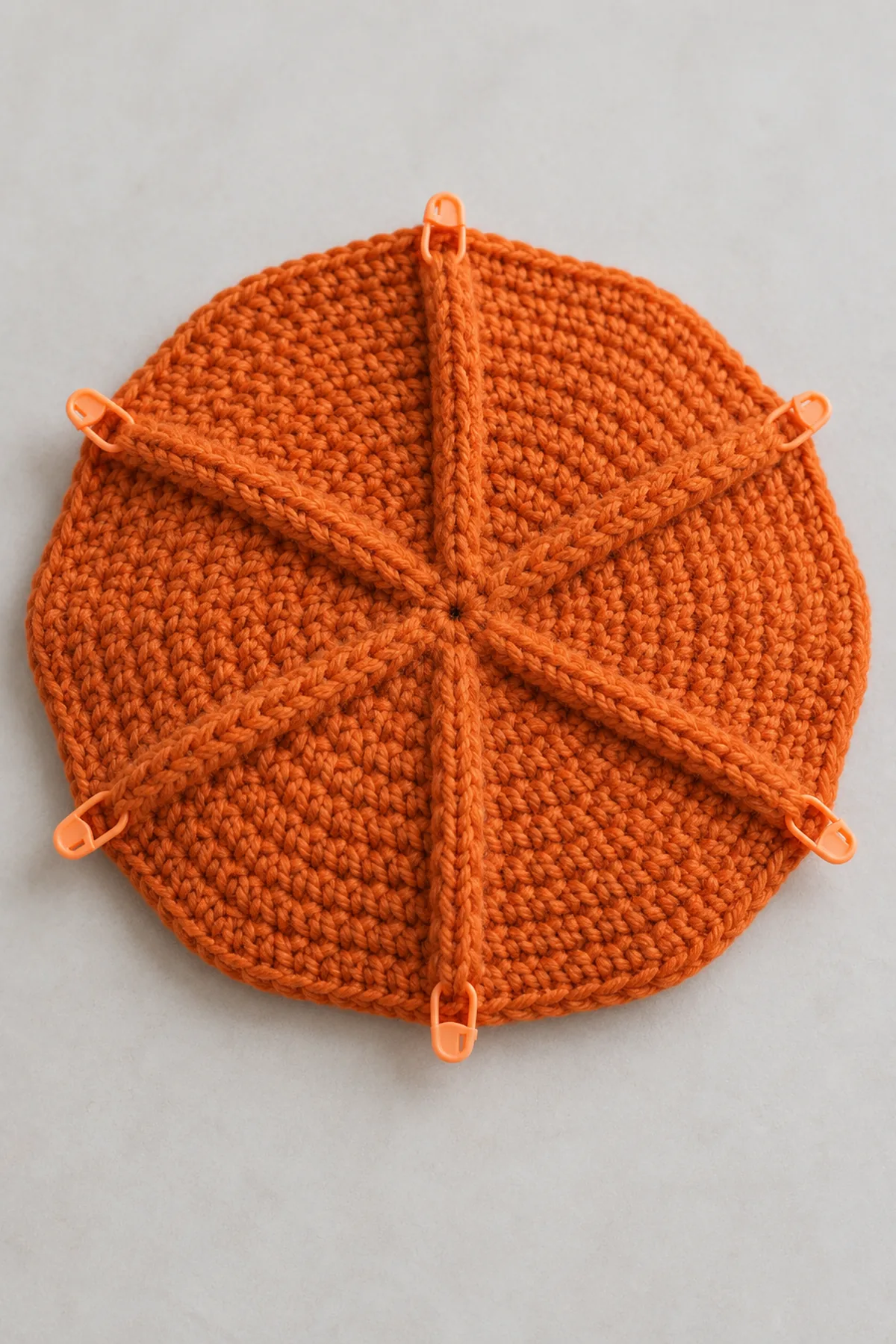 Partially grown broad orange crochet beret crown laid flat with six visible radial raised ribs and movable sector markers, no band or stem