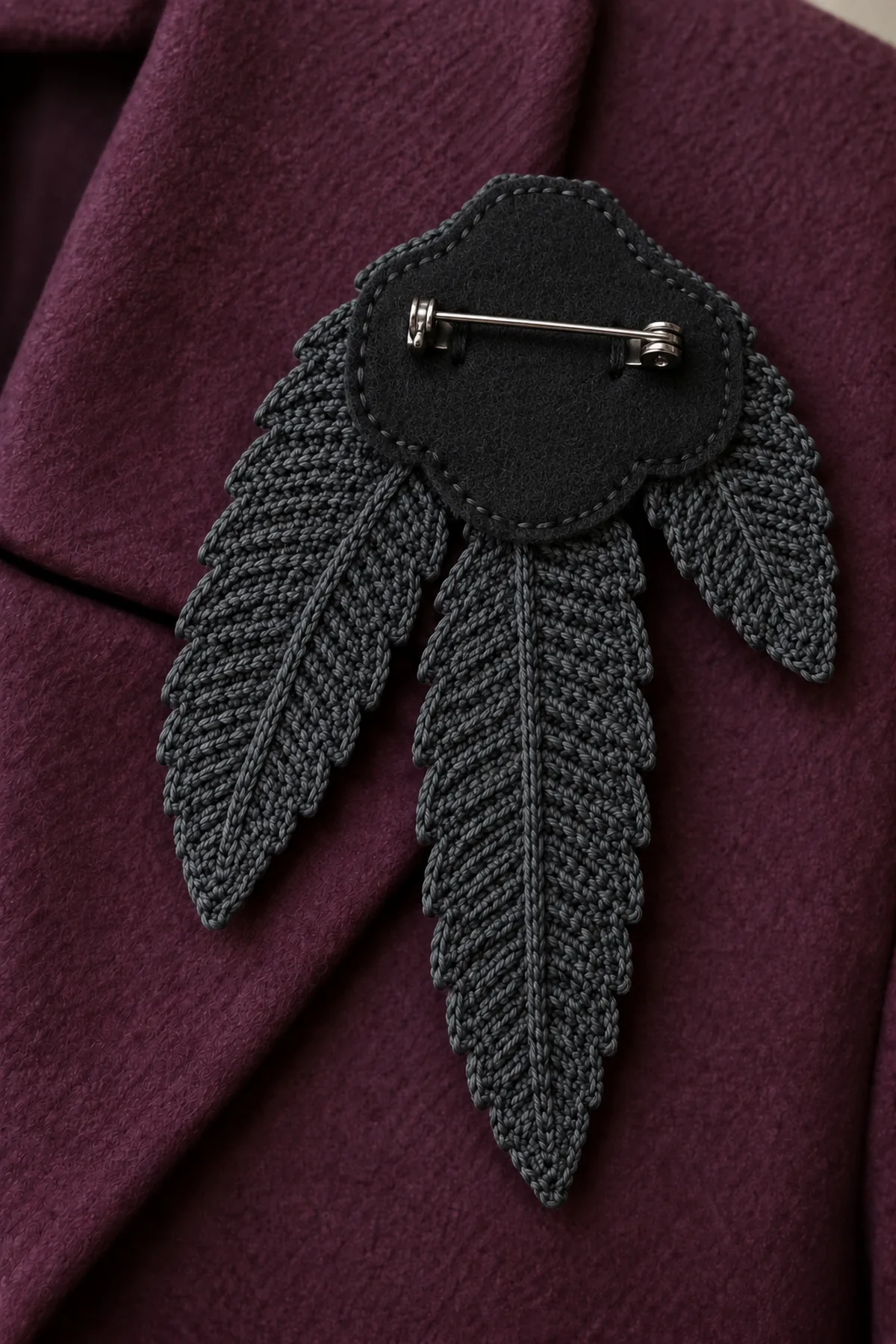 Reverse close-up of charcoal crochet three-feather brooch bases, two neatly stitched black felt layers with closed commercial locking pin bar sewn securely to outward felt face, sharp point enclosed in clasp, crochet tips extend away, no loose needles