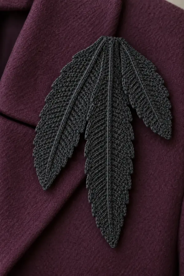 Original charcoal fine-cotton crochet raven feather shoulder brooch, THREE flat graduated tapered feather blades fanned downward from overlapping broad bases, subtle asymmetric stepped edges, central raised charcoal surface-slip-stitch rib on each. Longest center blade4.4in, medium left3.6in, small right2.9in. Hidden felt and locking pin behind bases. Laid on plum coat fabric, no real feathers, beads, fringe, face, wire or person