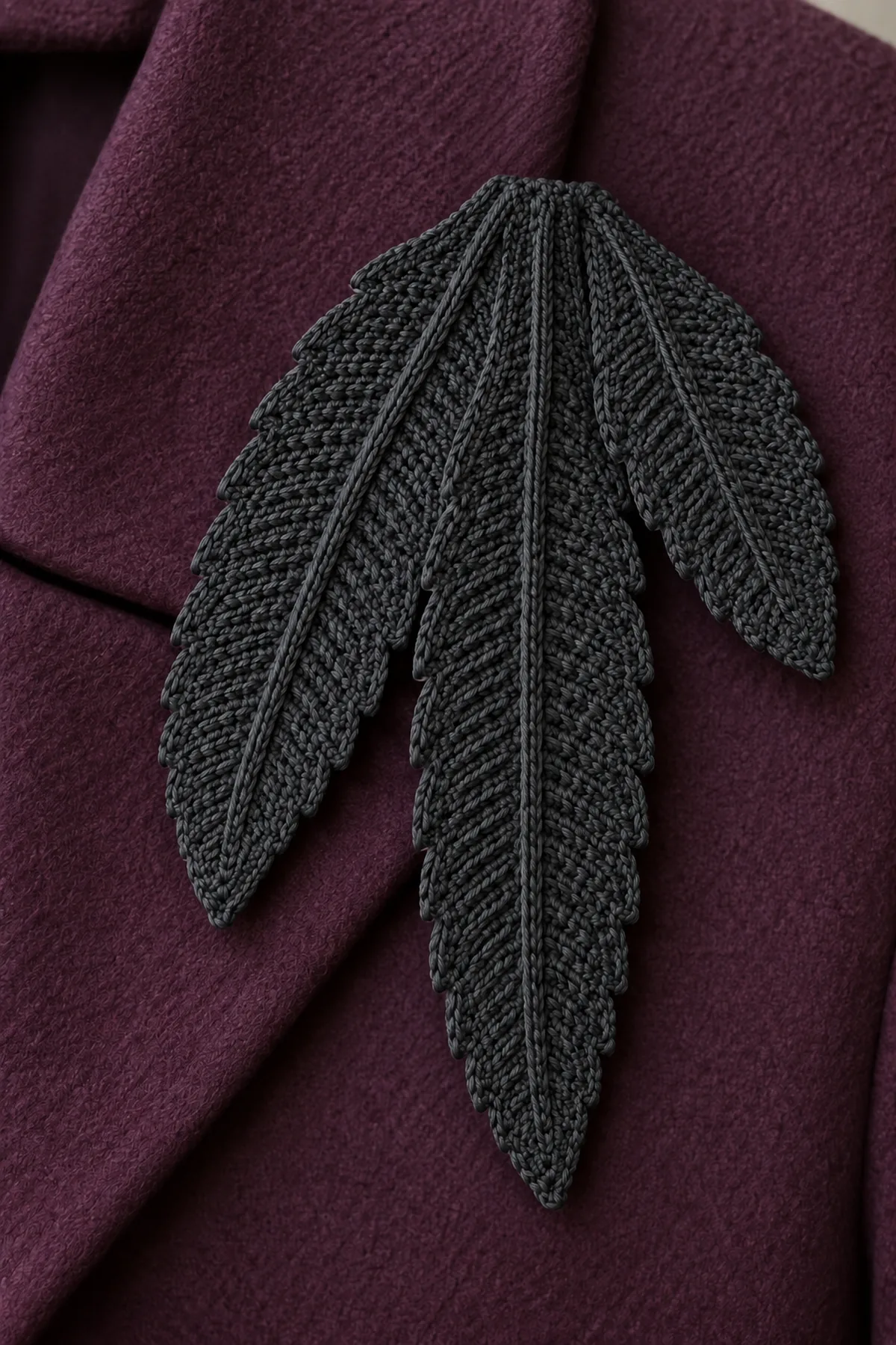 Original charcoal fine-cotton crochet raven feather shoulder brooch, THREE flat graduated tapered feather blades fanned downward from overlapping broad bases, subtle asymmetric stepped edges, central raised charcoal surface-slip-stitch rib on each. Longest center blade4.4in, medium left3.6in, small right2.9in. Hidden felt and locking pin behind bases. Laid on plum coat fabric, no real feathers, beads, fringe, face, wire or person
