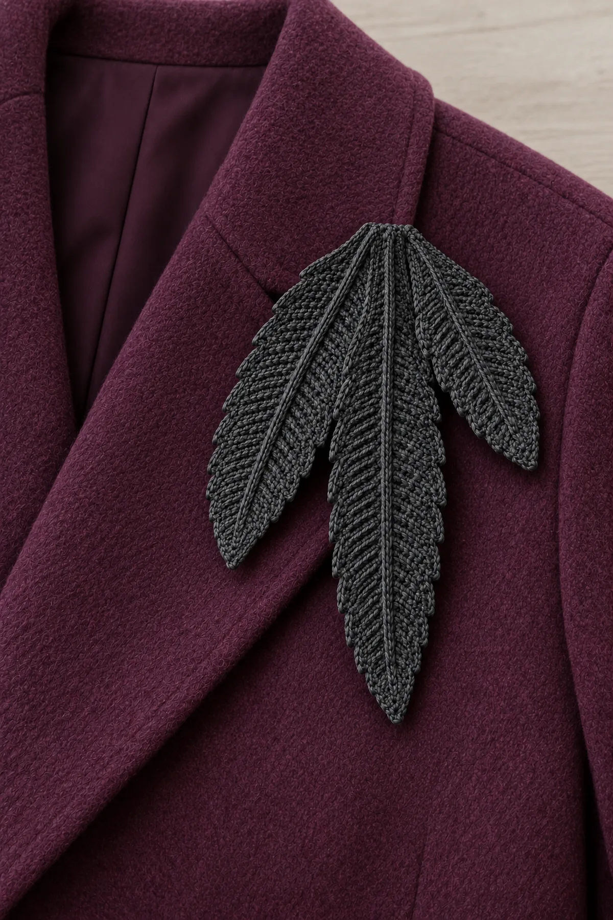 Same three graduated charcoal crochet feather blades with raised central ribs and stepped edges, brooch pinned on shoulder area of an unworn plum adult coat laid flat, longest feather points down, hidden pin backing, no person, jewelry or additional feathers