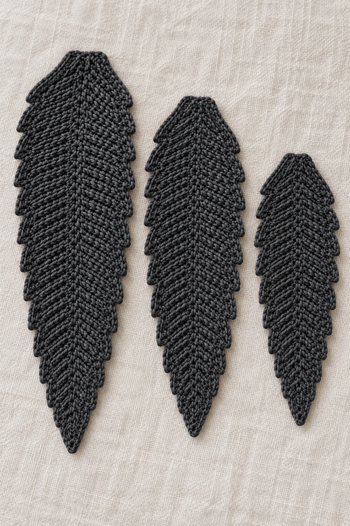 Three separate flat graduated charcoal crochet feather blades with tapered tips and subtle stepped edges, laid side by side on pale linen before adding central surface ribs or assembly, no real feather barbs, no felt attached