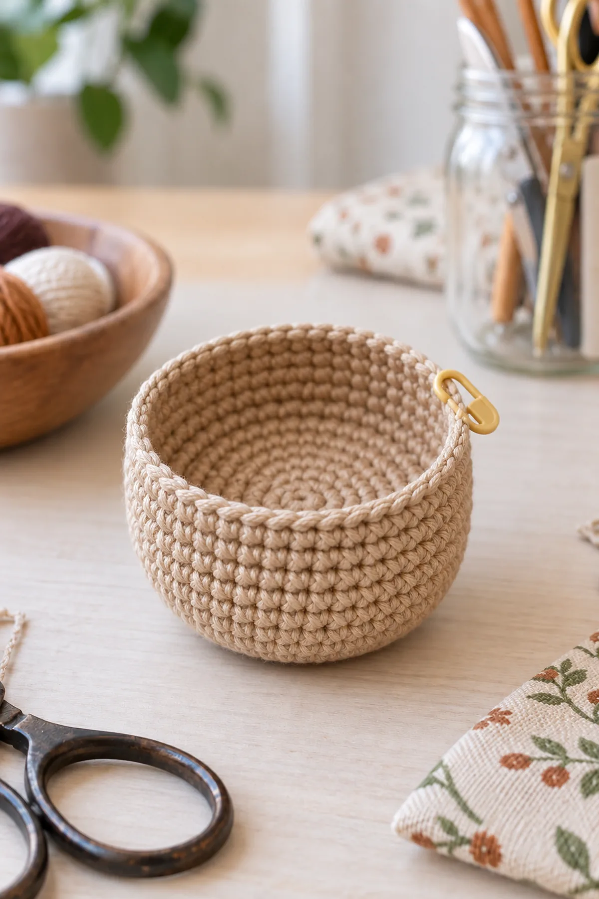 Small tan crochet flat-base cup with straight short walls and fully open top, empty, stitch marker at rim; broad circular bottom visible angled toward camera. No cocoa cap, no filling, no eyes. Pale sewing table