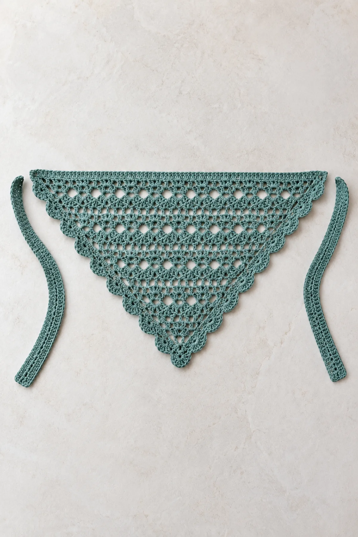 Completed shallow muted-teal lace triangle laid flat with scalloped edging along both sloping sides, separate broad crocheted ties not yet sewn on