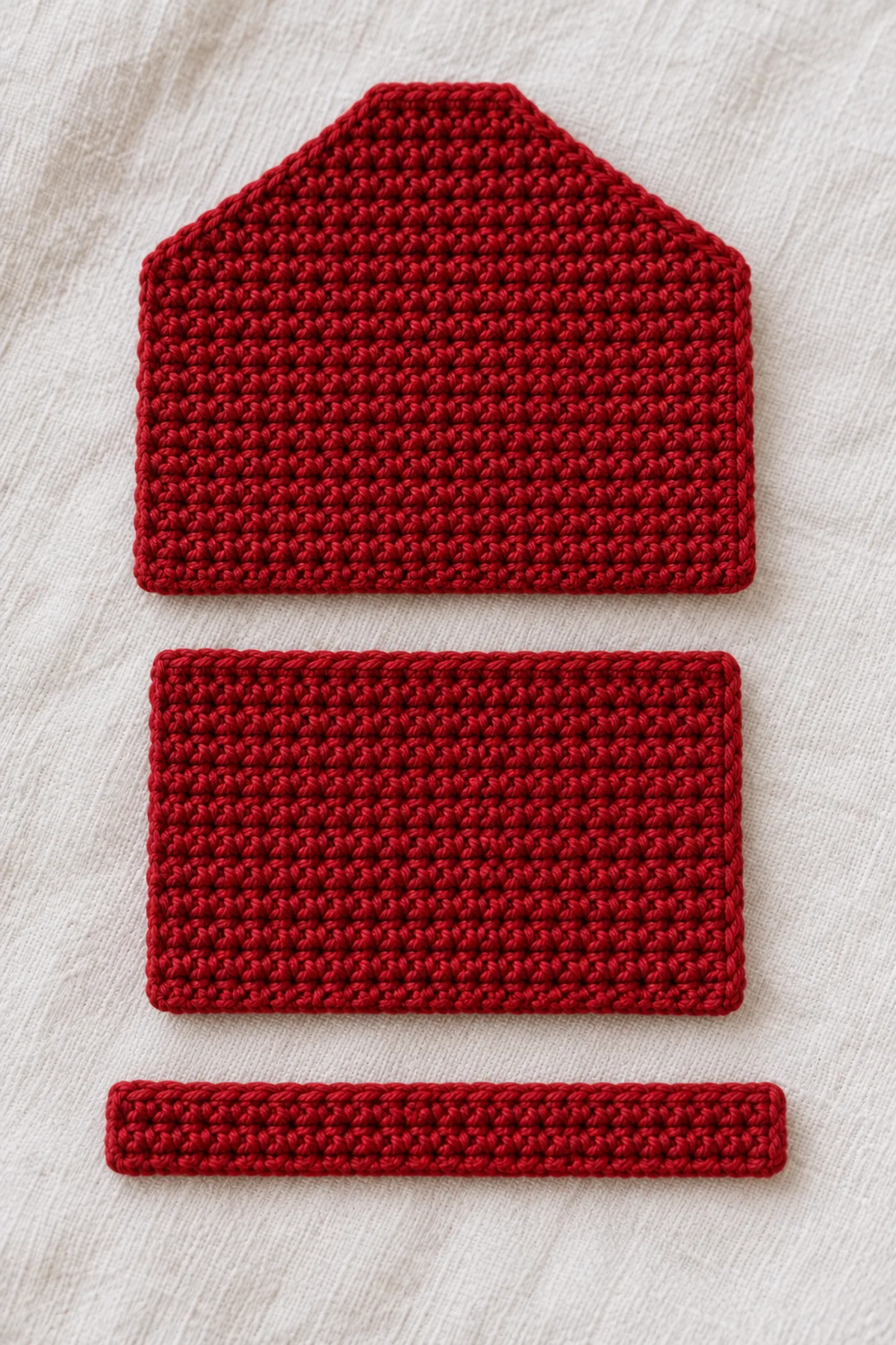 Two separate cranberry crochet wallet panels laid flat, one short rectangular front and one longer back continuing into a tapered six-stitch flap tip, a narrow red keeper strip beside them, no assembled wallet yet