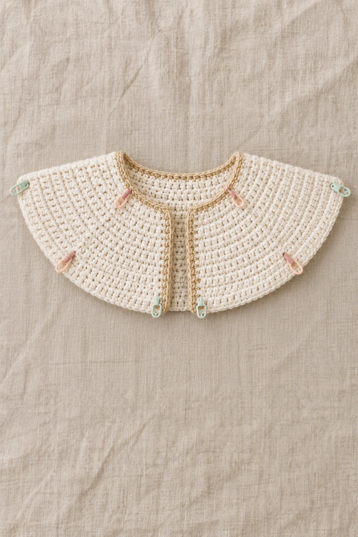 Partly worked cream open baby cardigan yoke laid flat in a broad gently curved arc; seven evenly distributed increase rows visible with multiple small removable group markers, no sleeves or body yet.