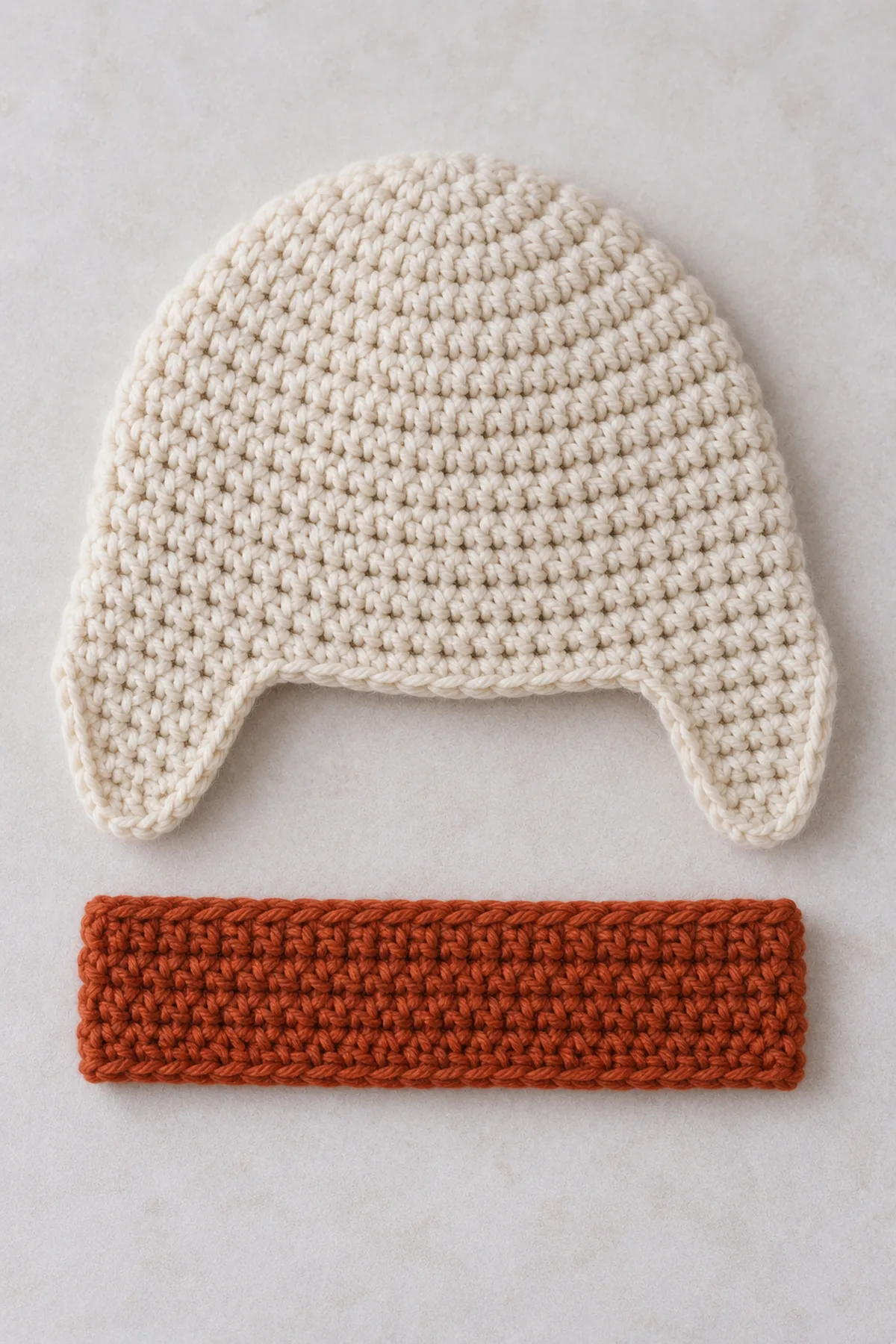 Cream fitted crochet cap laid flat with two short tapered integral side earflaps worked, separate flat rectangular rust forehead band beside it, back edge not yet turned up.