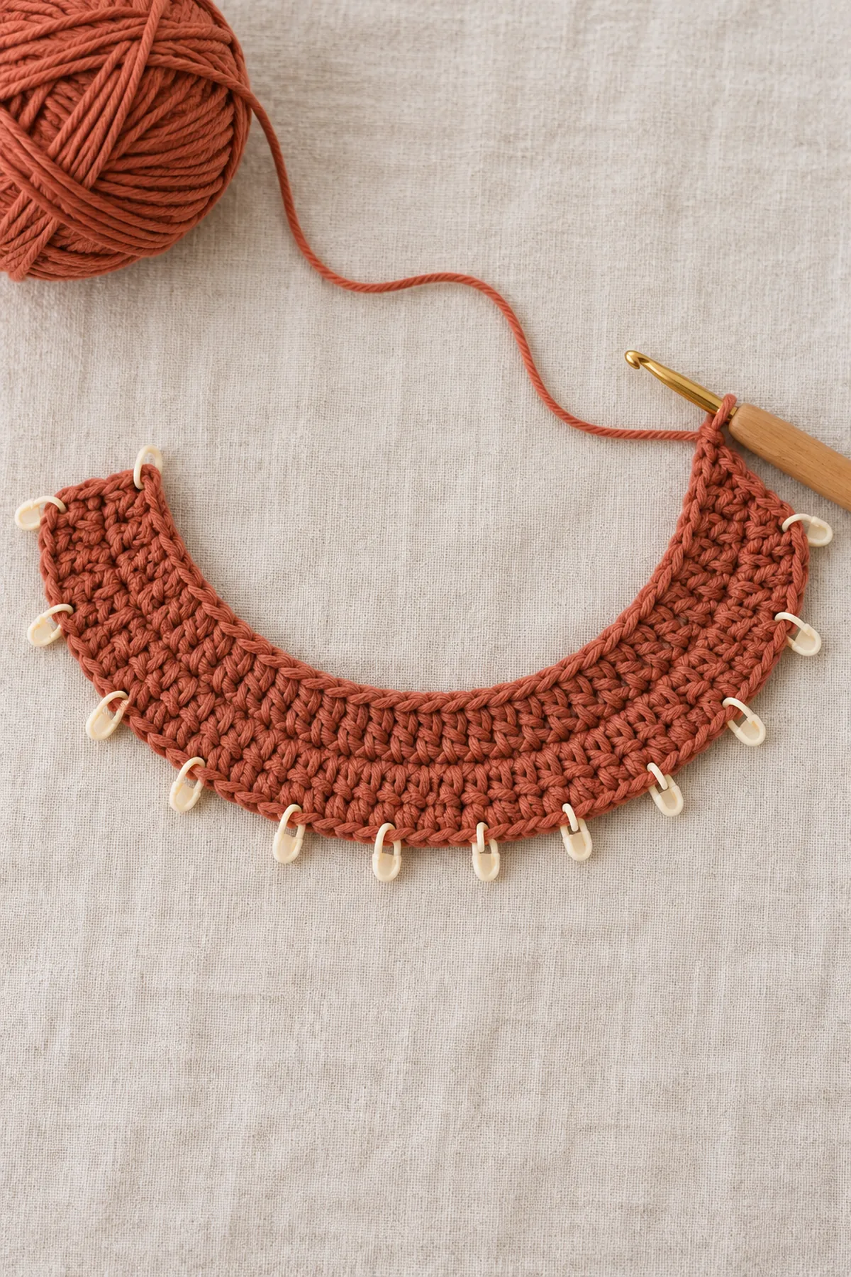 Open flat terracotta crochet neckline strip and three widening hdc rows forming shallow crescent, twelve evenly spaced markers on outer edge, working hook attached; no collar or tabs.