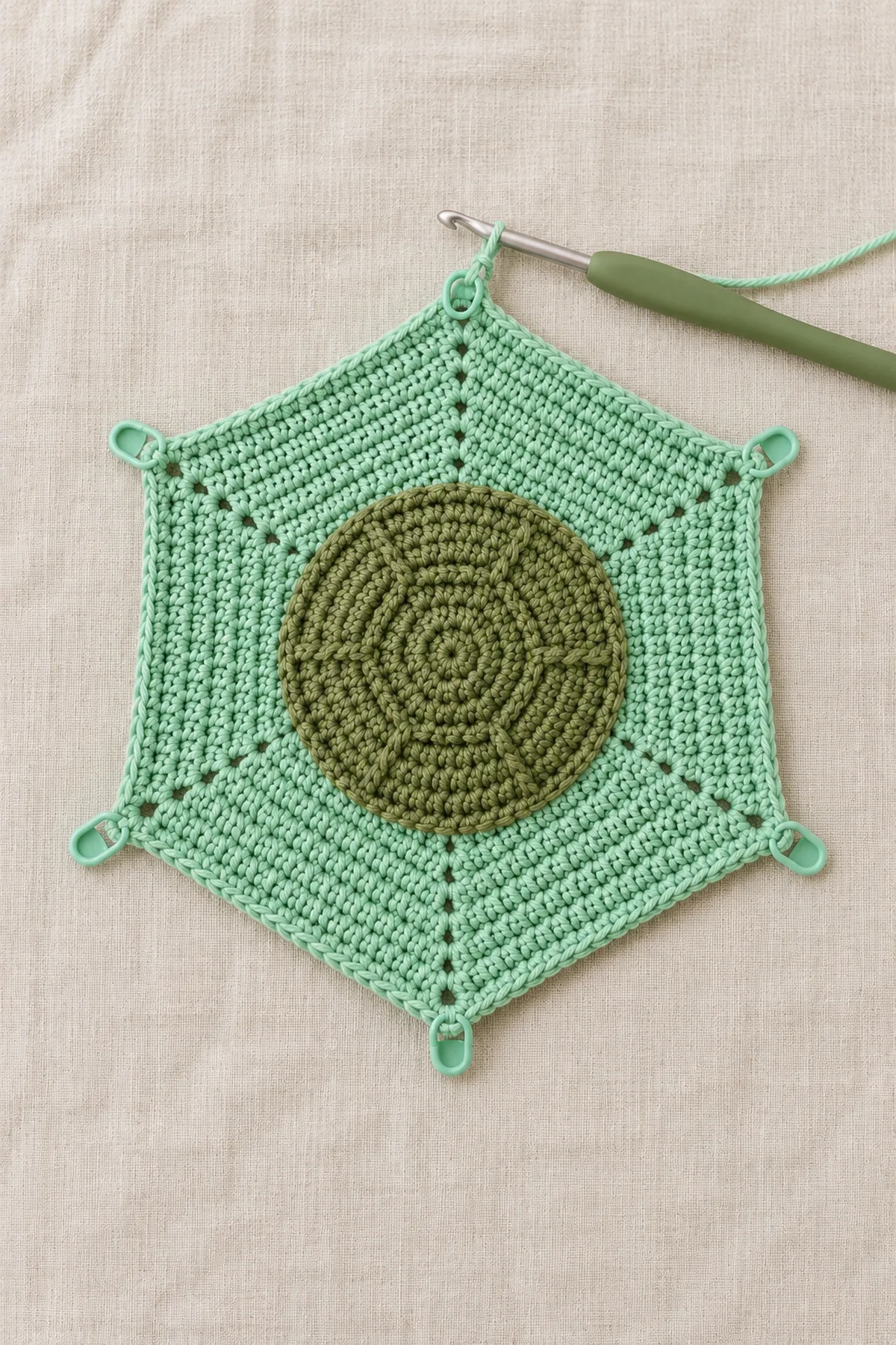 Mint six-sided flat crochet lovey cloth partway through center-out growth, six markers at corners, 12 visible rounds, hook attached; no shell, head or flippers.