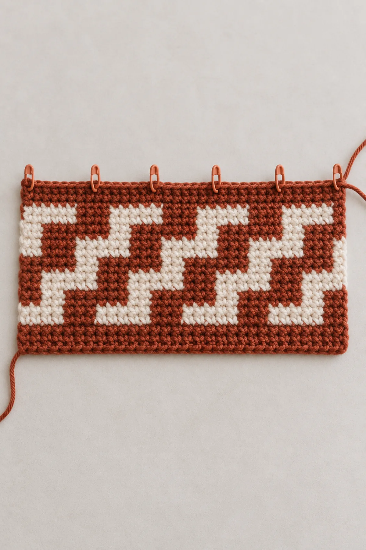 Flat partially worked rust-and-cream crochet overlay mosaic panel, right side up, early four-column staircase blocks visible and six repeat markers across width