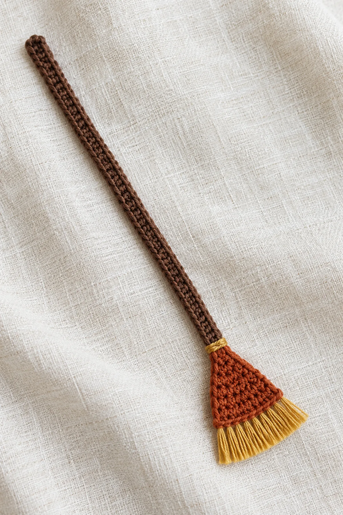 One thin flat crochet witch broom bookmark lying diagonally on ivory linen: long narrow cocoa single-crochet shaft with central raised cocoa slip-stitch line, tiny flared rust trapezoid crochet broom head, small gold band at shaft-head join, short straight gold cotton tassel bristles along broad end. No wooden stick, wire, stuffing, eyes or charm; about 7.6 inches long